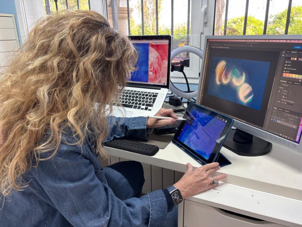 Mujer trabajando en un ordenador y tableta digital, rodeada de pantallas.